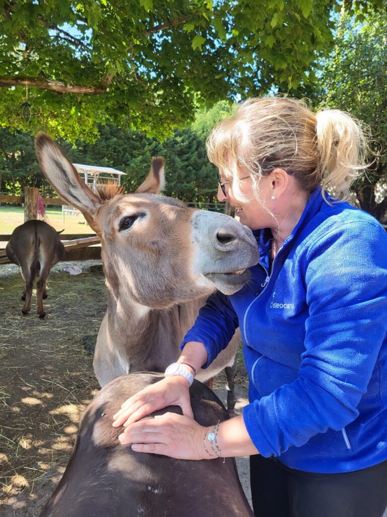 Sylvie Praz, enseignante et ostéopathe pour animaux, face à un âne gris, les mains posées sur le dos d'un second âne